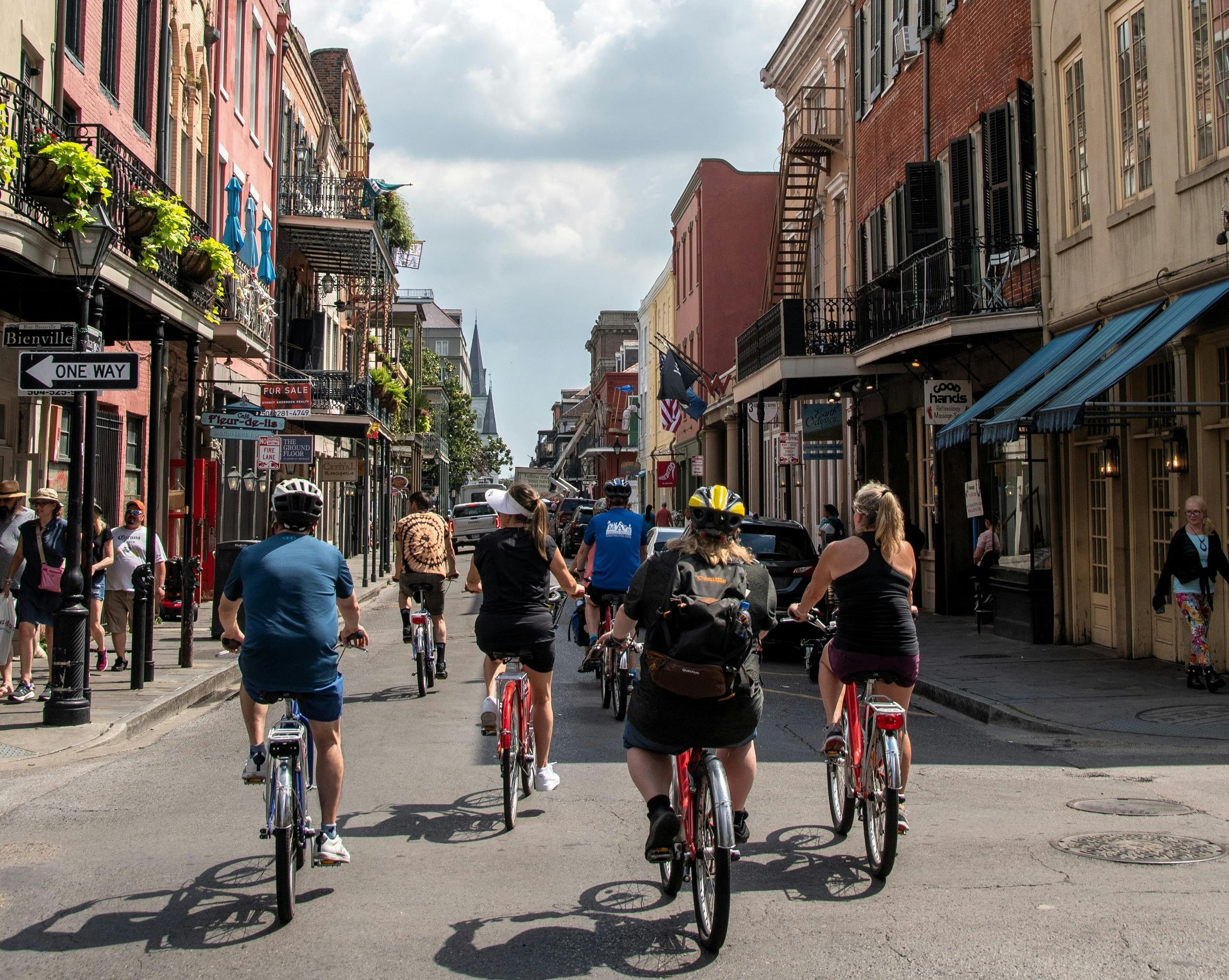 En gruppe cyklister kører ned ad en livlig gade i byen med historiske bygninger og butikker under en delvist overskyet himmel.
