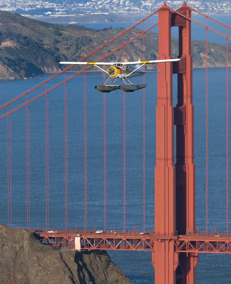 A seaplane flies in front of the Golden Gate Bridge with the ocean and distant hills in the background.