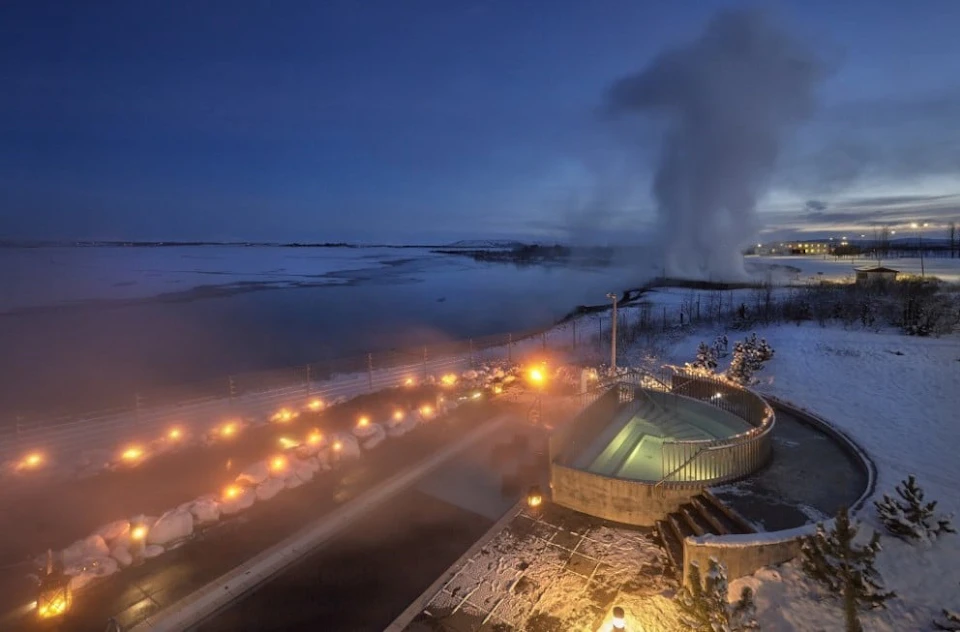 Outdoor hot spring pool at dusk with steam rising, surrounded by snow, illuminated by warm lights under a dark blue sky.