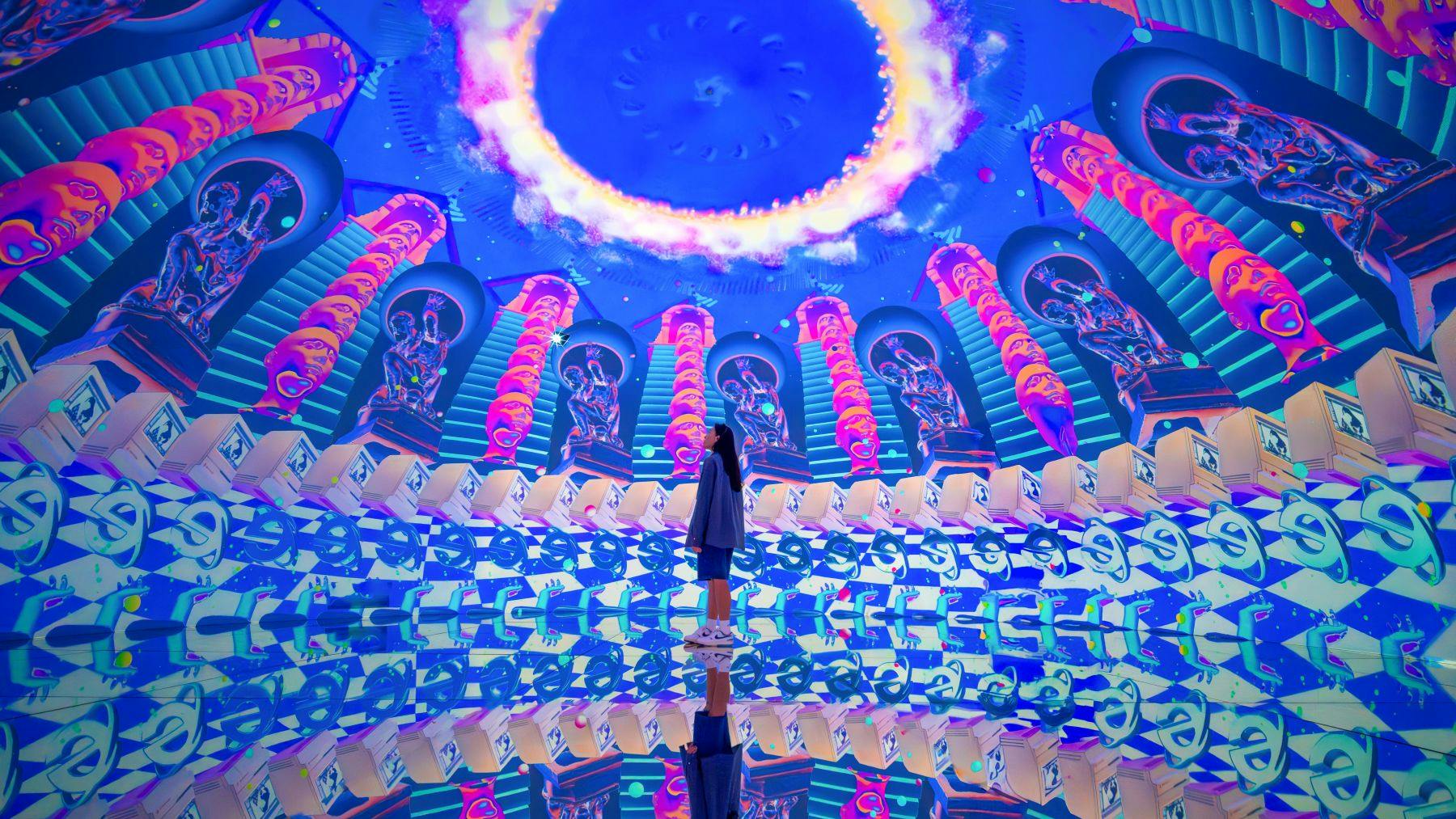 Eine Person steht in einer farbenfrohen, immersiven Kuppel mit lebhaften, symmetrischen Mustern und Reflexionen auf dem Boden.