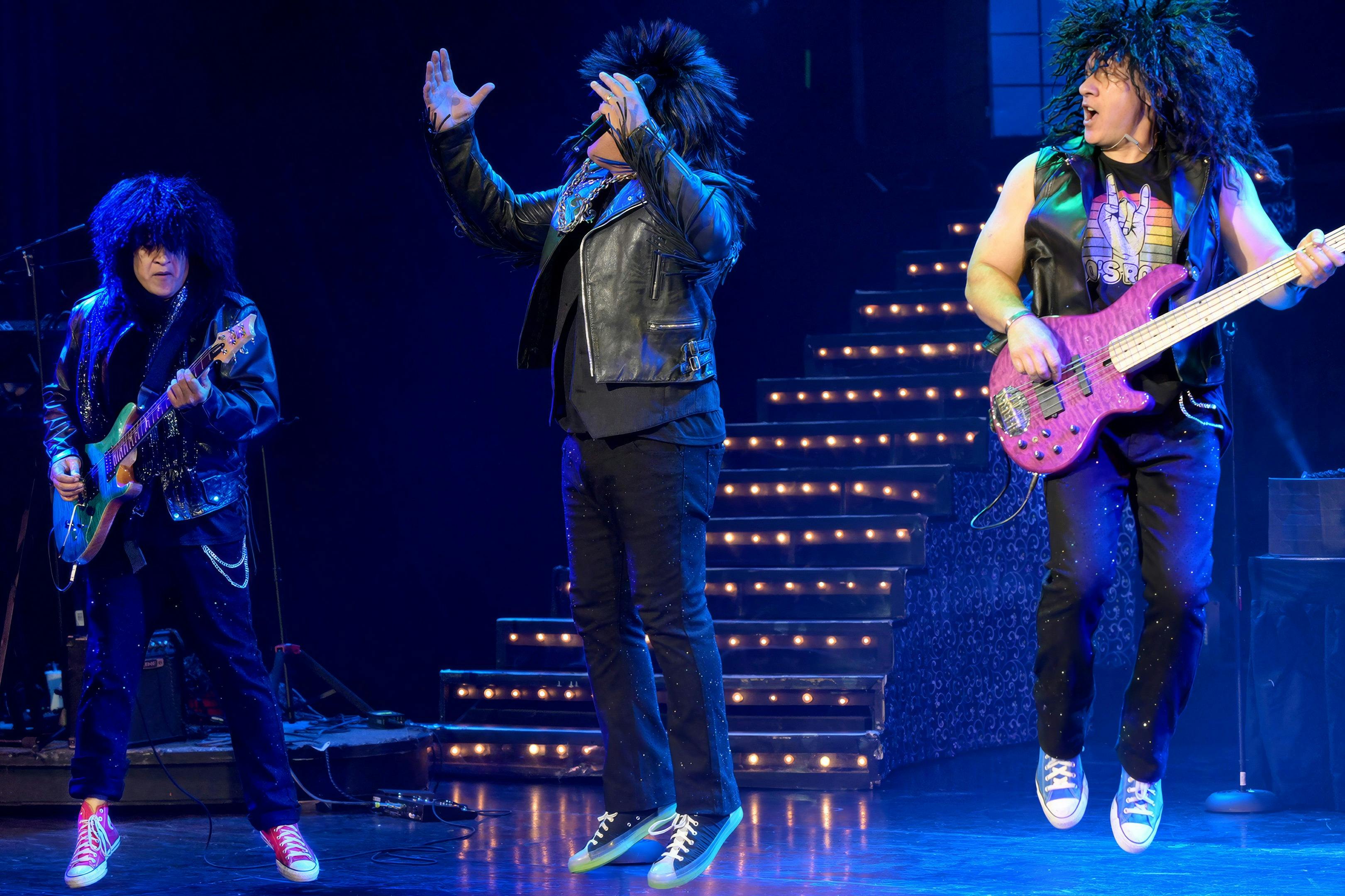 Two performers on stage wearing wigs and costumes, one playing a pink bass guitar, with lit stairs in the background.
