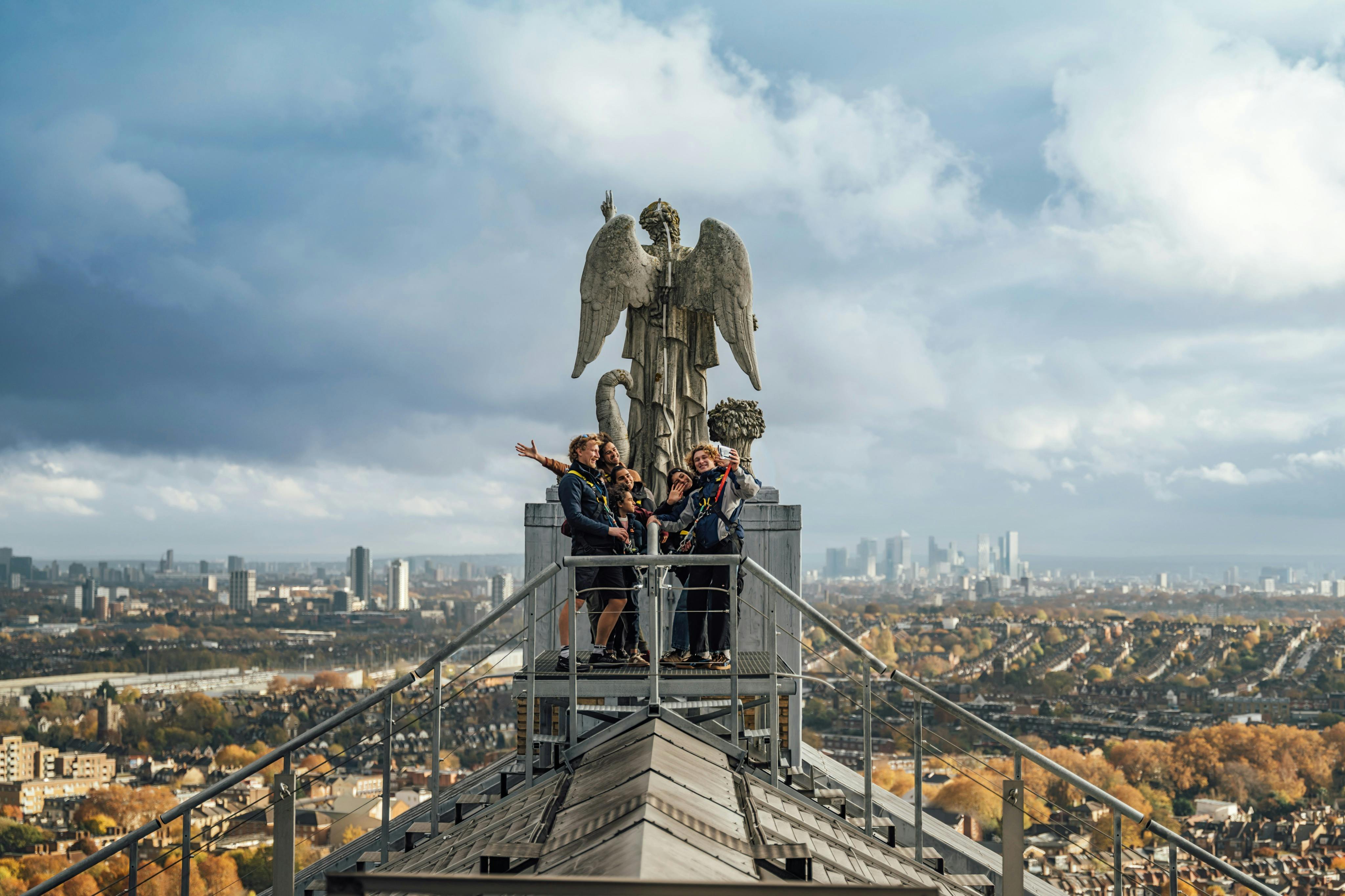 A group of people standing on a rooftop platform near a statue of an angel, with a cityscape in the background.