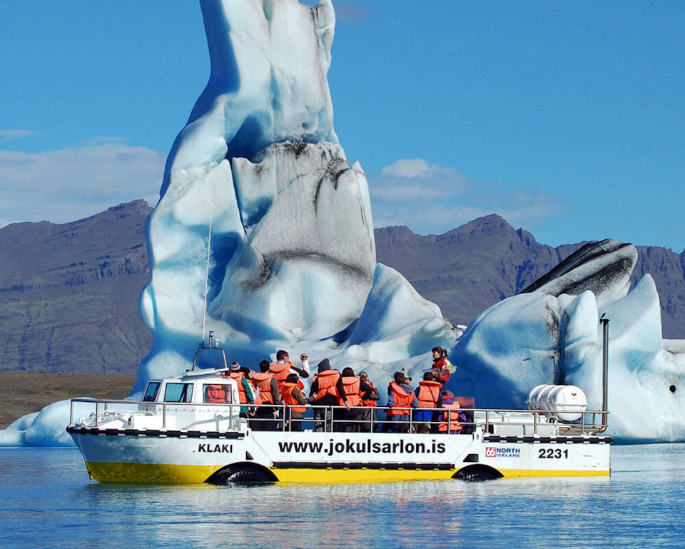Tourists in orange life jackets on a boat near a large iceberg in a serene body of water with mountains in the background.
