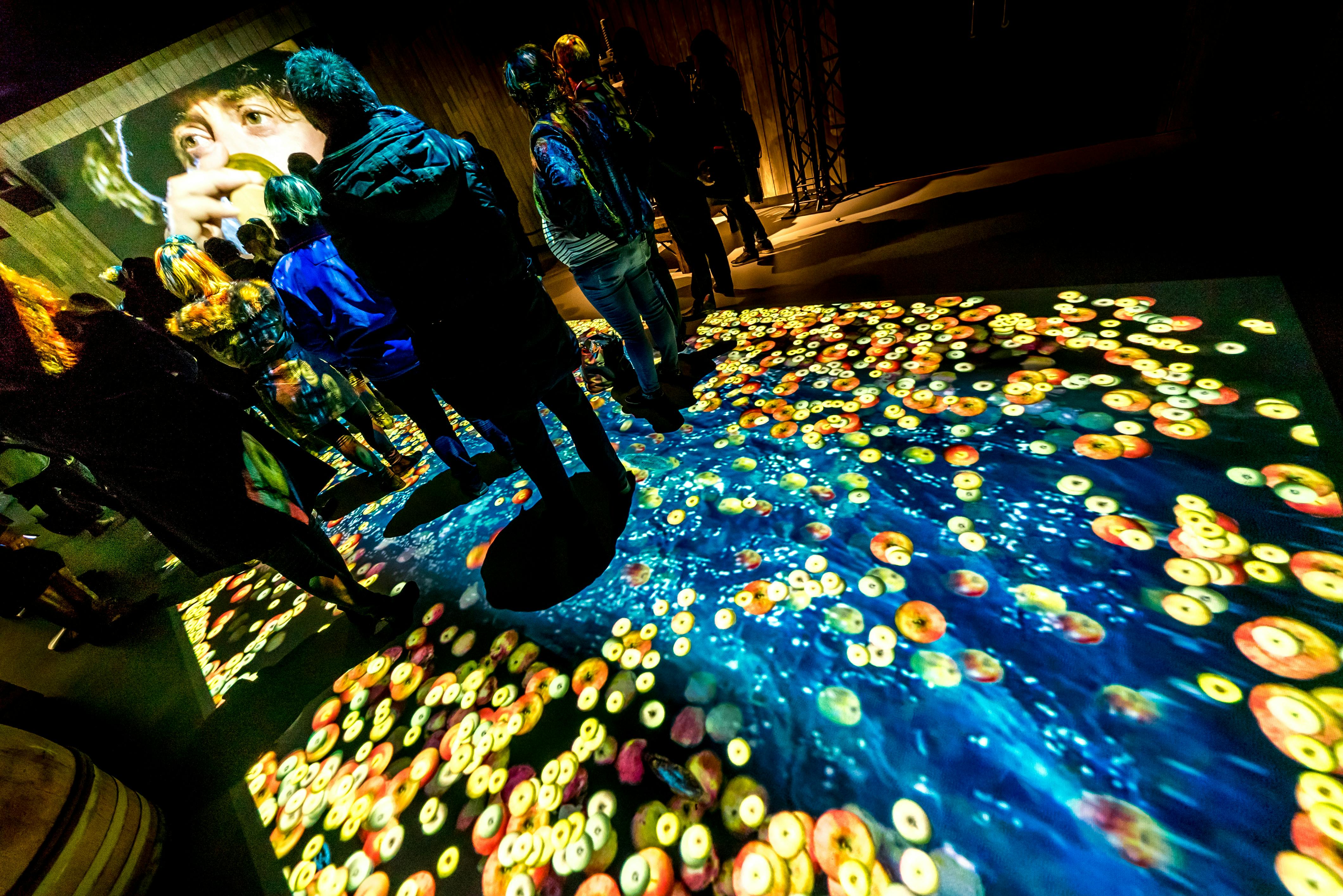 La gent s'aixeca sobre un terra i està envoltada de parets amb imatges artístiques projectades i acolorides, incloent-hi un gran rostre i patrons abstractes.