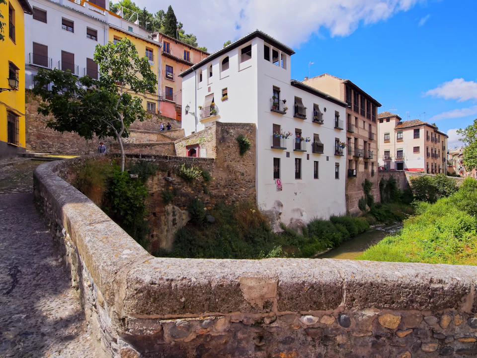 A stone bridge overlooks a river with multi-story buildings and greenery in the background under a partly cloudy sky.