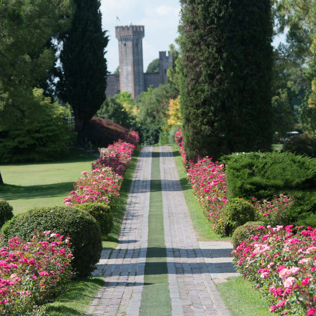 A paved pathway lined with pink flowers and tall trees leads to a castle in the background on a sunny day.