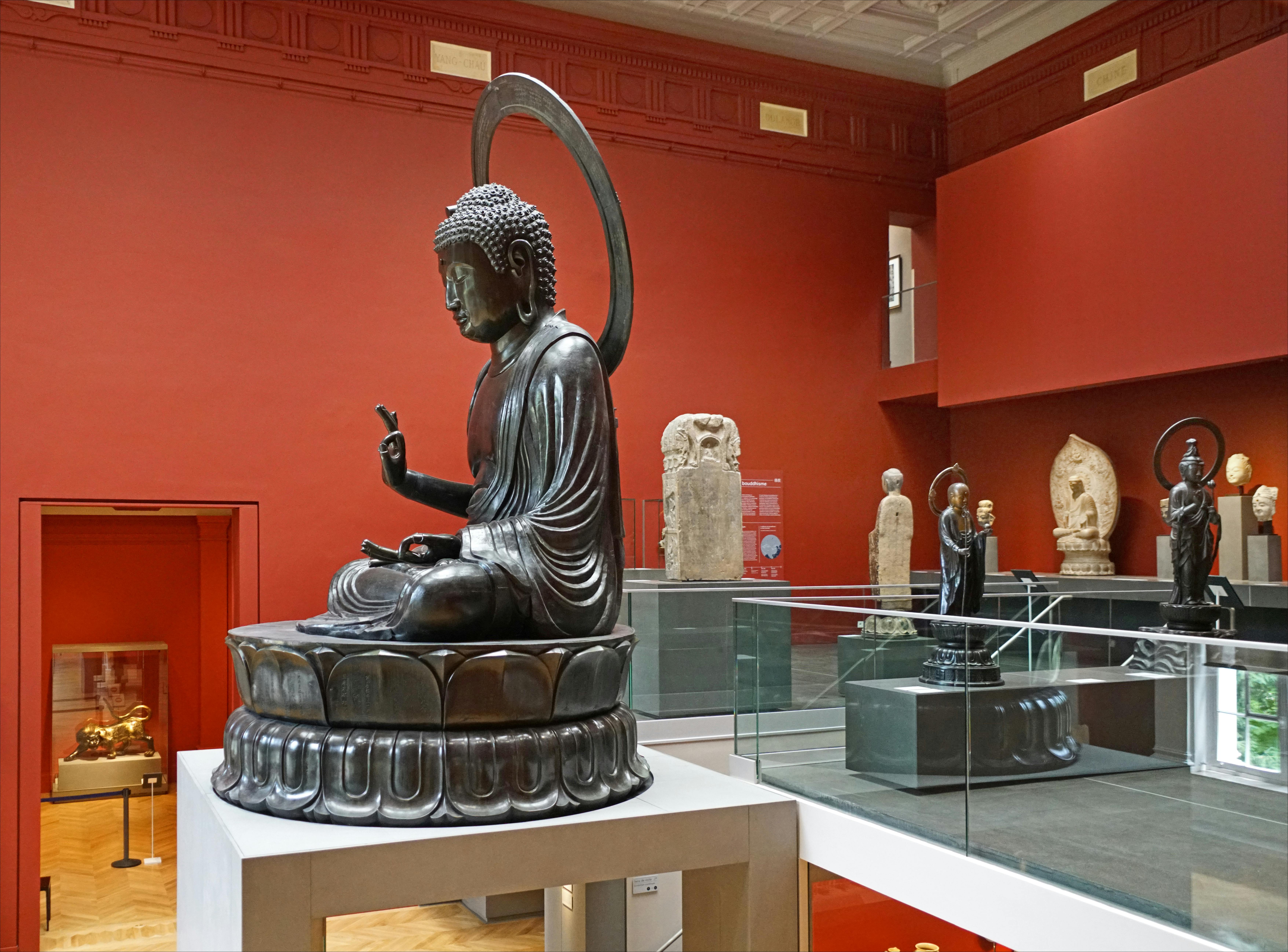 博物馆底座上有一尊大型青铜佛像，背景是红墙和各种其他雕塑。