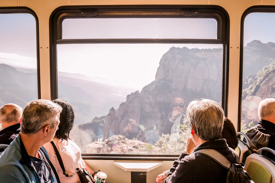 Three passengers look out a large train window at a mountainous landscape with cliffs and valleys. Reflections are visible.