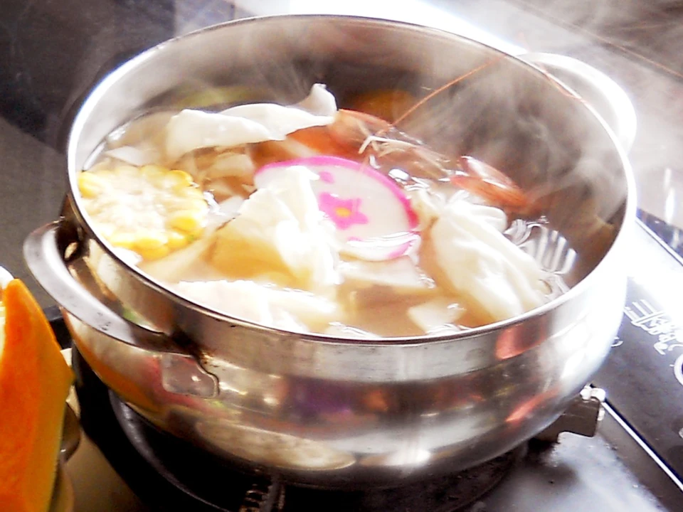 A meal setup featuring a hot pot on a portable stove, raw ingredients, a bowl of rice, a soft-boiled egg, and condiments.