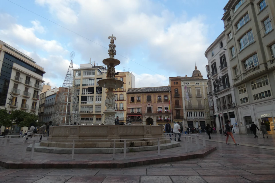 Town square with a central fountain, surrounding buildings with balconies, and people walking. A metal framework tree structure is present.