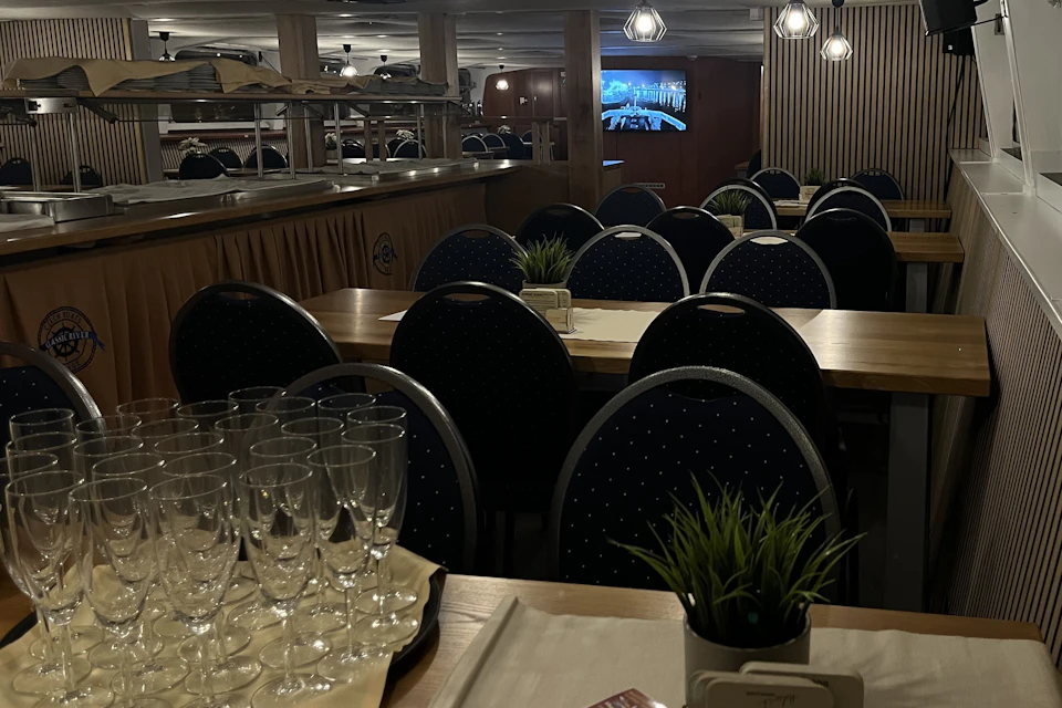 A dining area with tables set with glasses and potted plants, dark chairs, and a screen visible in the background.