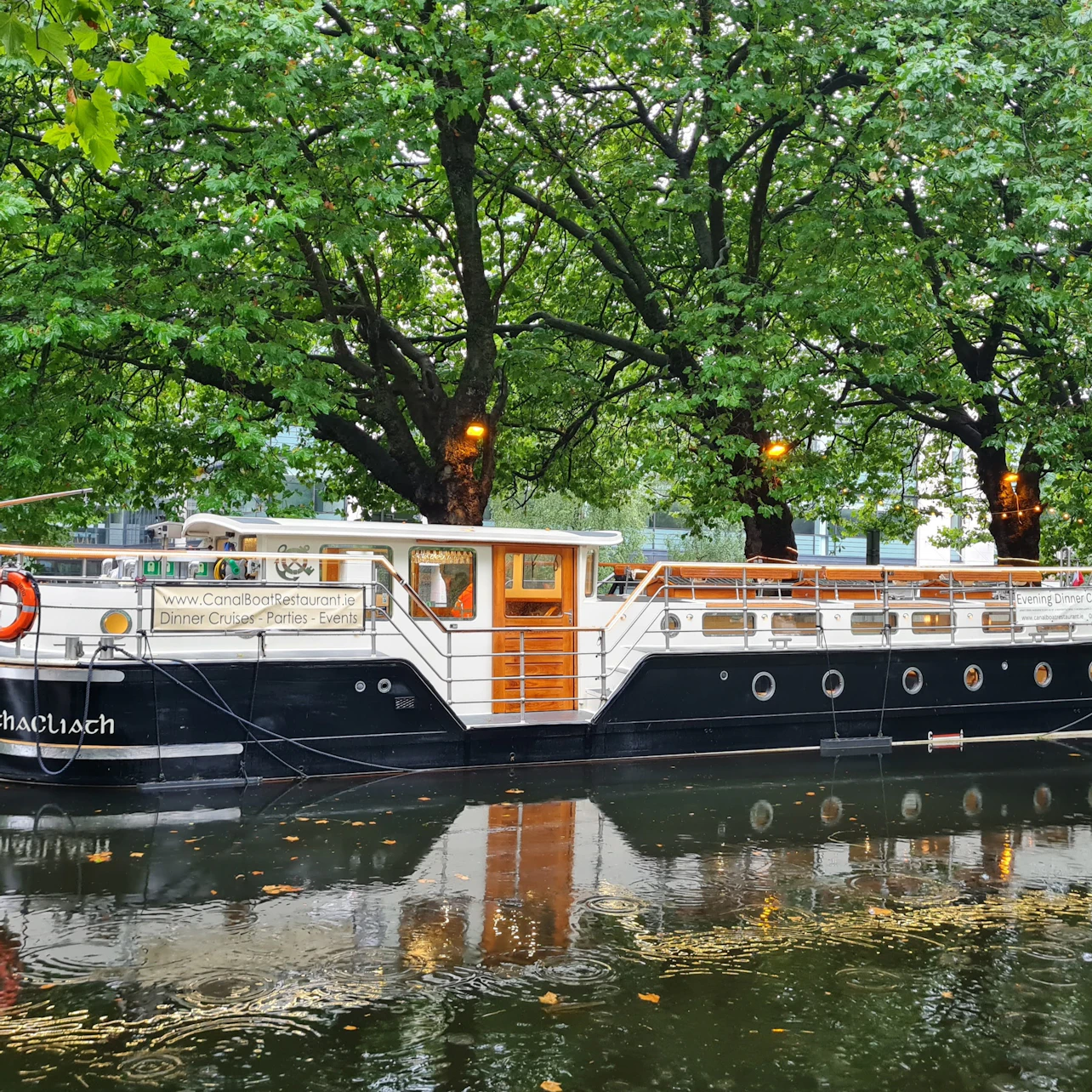 A moored canal boat with black and white exterior and wooden details, set against lush green trees and calm water.