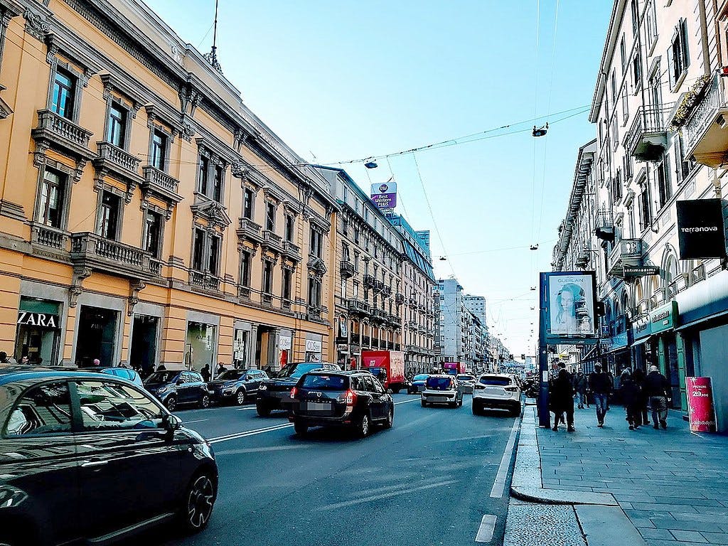 Urban street with traffic, people walking on sidewalks, and storefronts including Zara. Historic buildings line both sides under a clear sky.