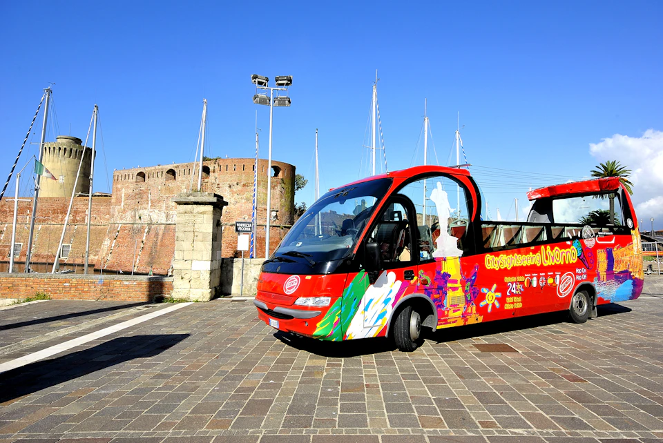 A vibrant red sightseeing bus near an ancient stone fortress with sailboat masts rising in the background on a sunny day.