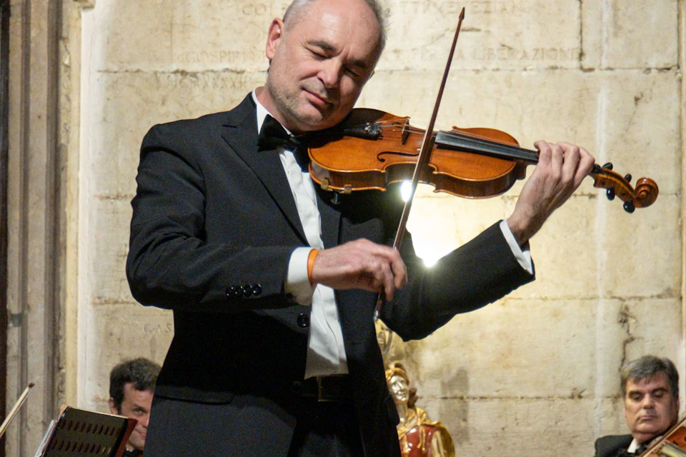 A string ensemble of six musicians performing indoors, with one standing violinist and five seated players in formal attire.