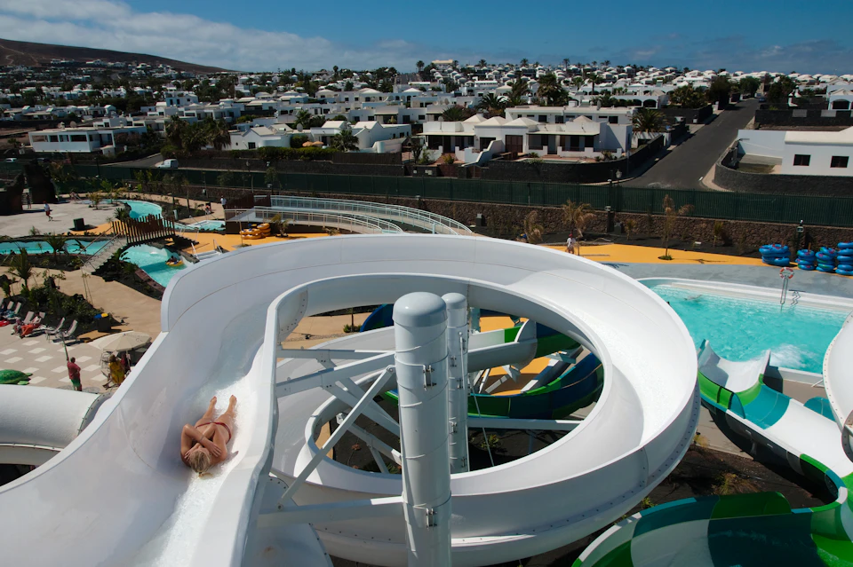 A person sliding down a white twisty water slide at a water park with rows of white houses in the background.