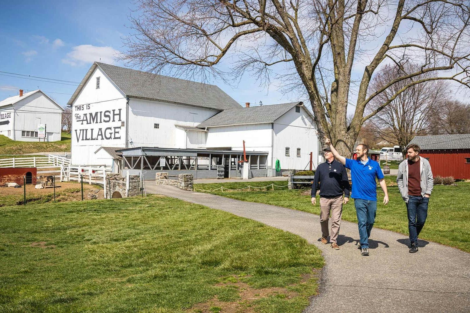 Three men walking on a paved path through a grassy area, with barns and leafless trees in the background on a sunny day.