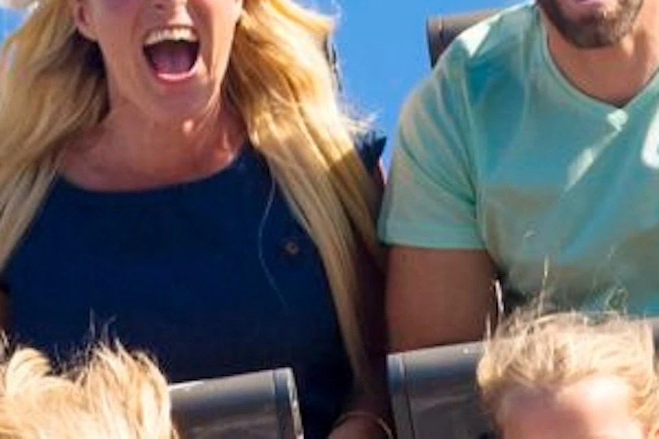 A family of four excitedly rides a rollercoaster with a yellow front, blue track, and a clear blue sky in the background.