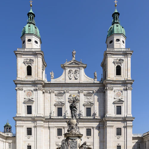 En stor barockkatedral med tvillingtorn, en central staty och människor som går på en torg under en klarblå himmel.