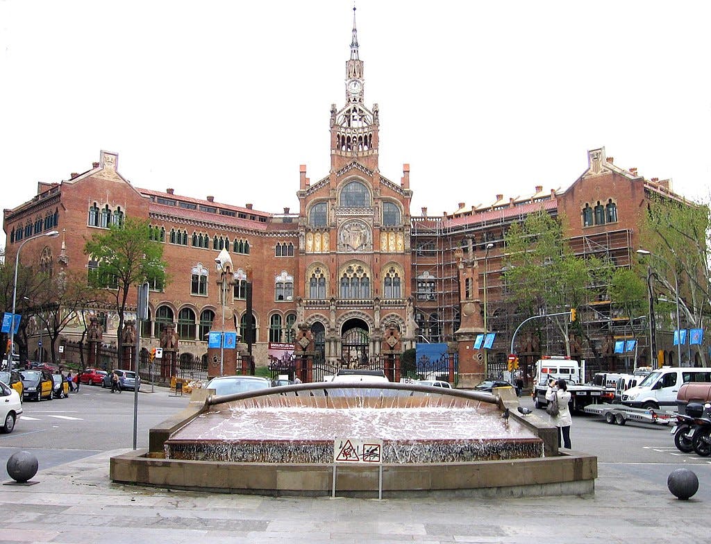 A historic building with a clock tower and ornate architecture, a foreground fountain, surrounding cars, trees, and scaffolding.