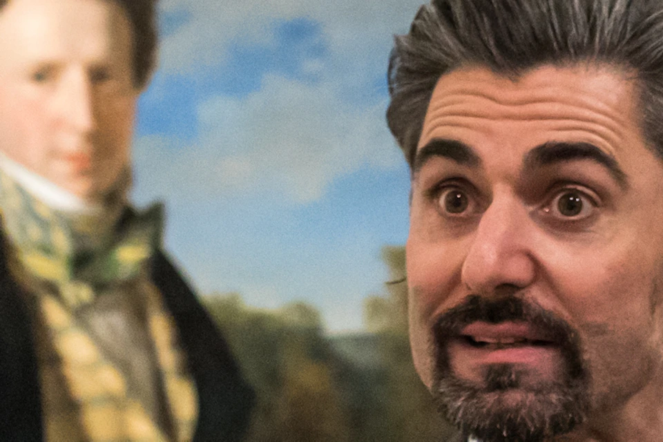 A man in a suit gestures while speaking to two men, with a portrait of a historical figure in the background.