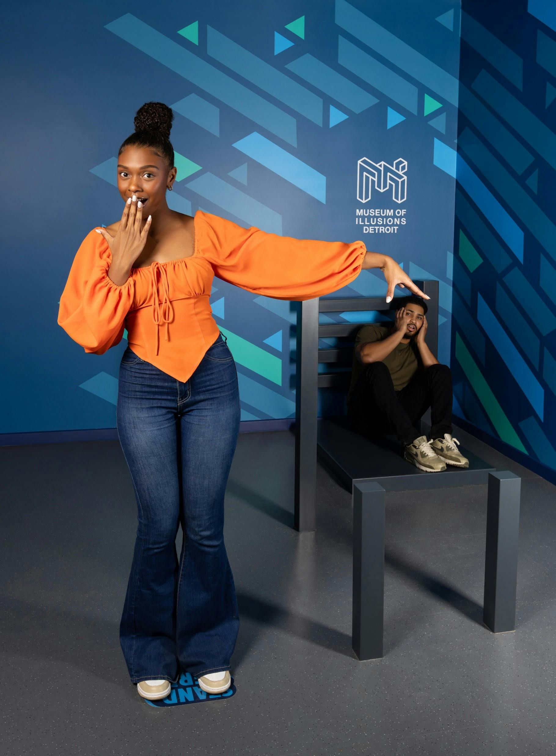 Μια γυναίκα με πορτοκαλί μπλούζα ποζάρει με το χέρι της να φαίνεται ότι αλληλεπιδρά με ένα άτομο που κάθεται σε μια μεγάλη καρέκλα σε μια διάταξη ψευδαίσθησης.