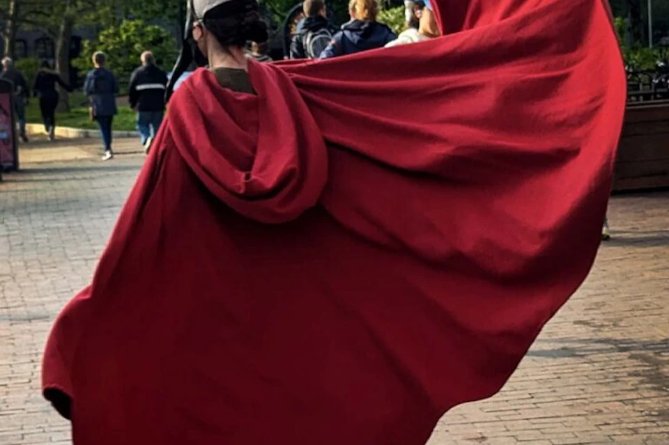 A person wearing a flowing red cloak outdoors, with other people visible in the background.