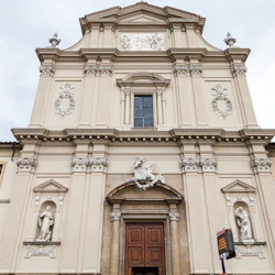 Fasad av en historisk beige kyrka med statyer i alkover, dekorativa stenarbeten och stora trädörrar under en molnig himmel.