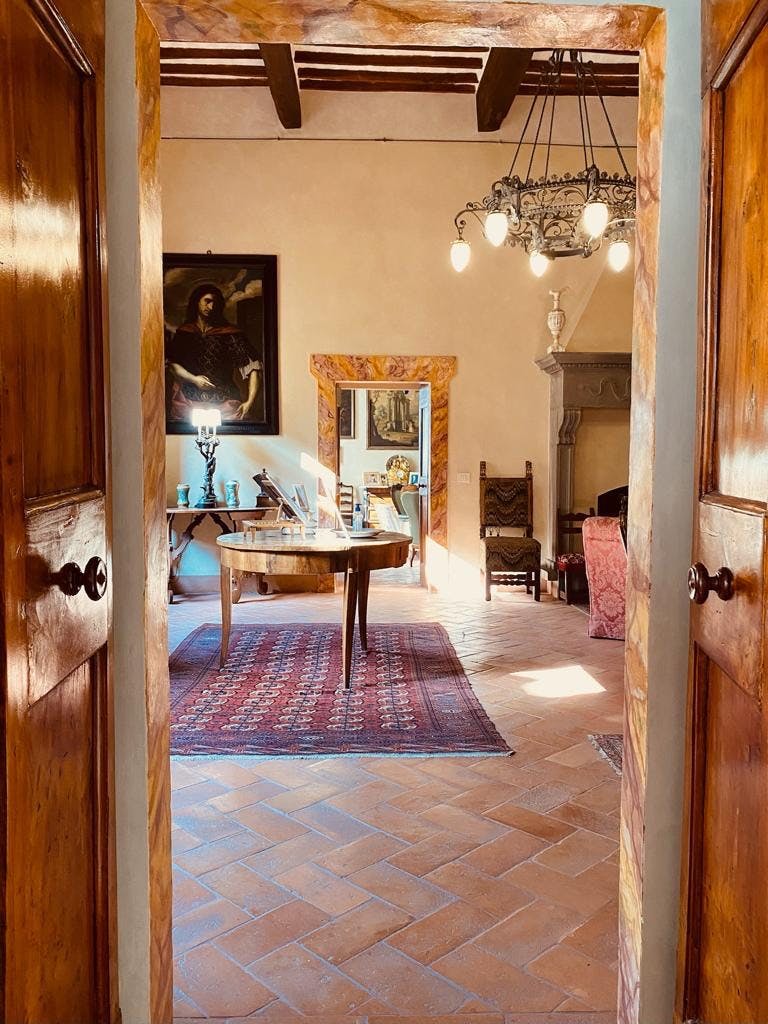 Una stanza caldamente illuminata con un tavolo da pranzo in legno su un tappeto rosso, un lampadario, un quadro, un camino e varie sedie e decorazioni.