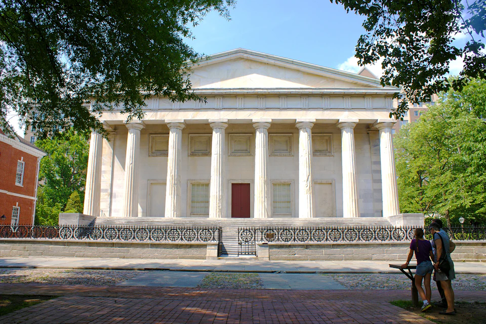 A neoclassical building with six columns at the front and a red door, surrounded by a decorative black wrought-iron fence.