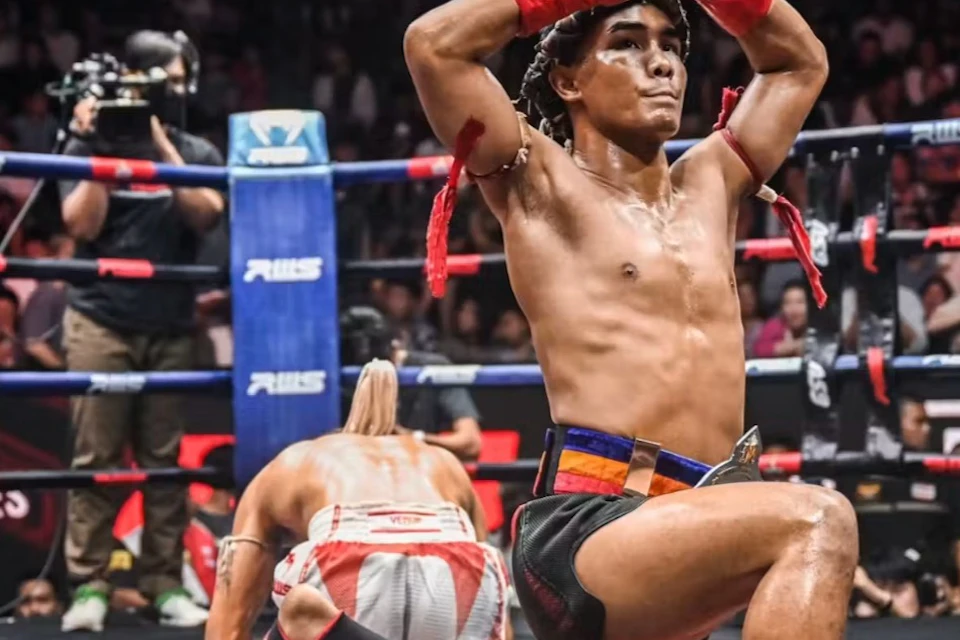 A martial artist kneels and performs a gesture in a boxing ring, with a crowd watching in the background.