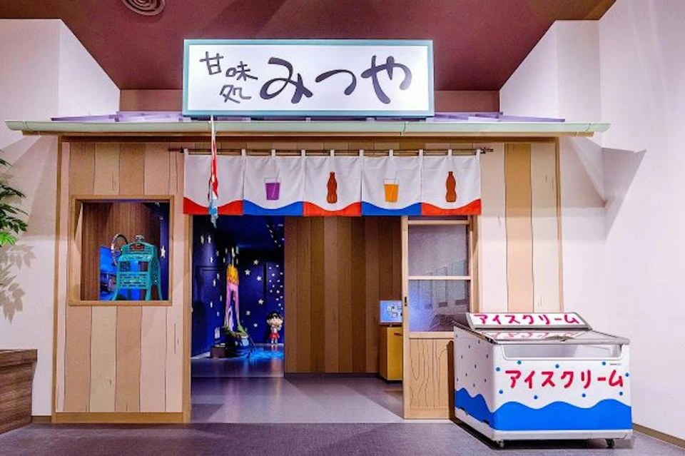 A Japanese-style storefront with a sign reading "みつや," featuring a colorful curtain and an ice cream freezer outside.