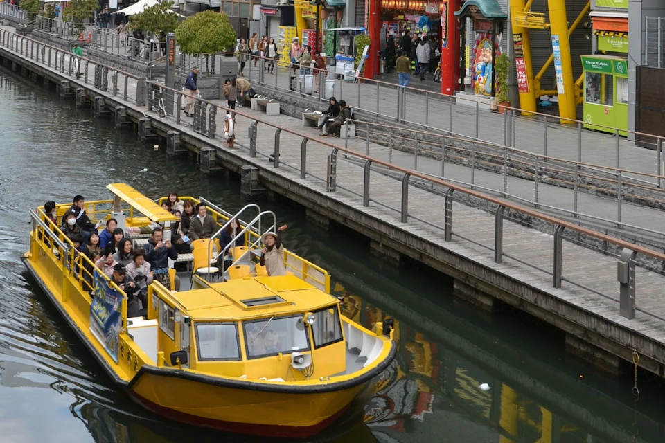 A yellow boat with passengers is cruising on a canal beside a promenade with shops and people walking.