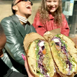 Two sandwiches held in the foreground and 3 women laughing in the background in front of a restaurant