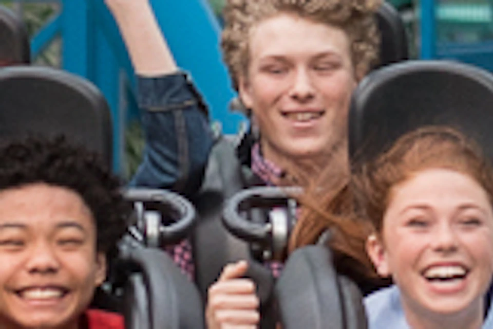 People riding a roller coaster with turquoise tracks inside an amusement park, with expressions of excitement and joy.
