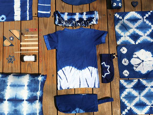 A collection of indigo-dyed items, including a t-shirt, pouches, and fabric pieces, arranged on a wooden surface.