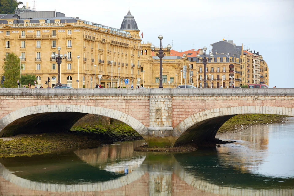 People walking across a stone bridge with arched openings over a calm river, with historic yellow buildings in the background.