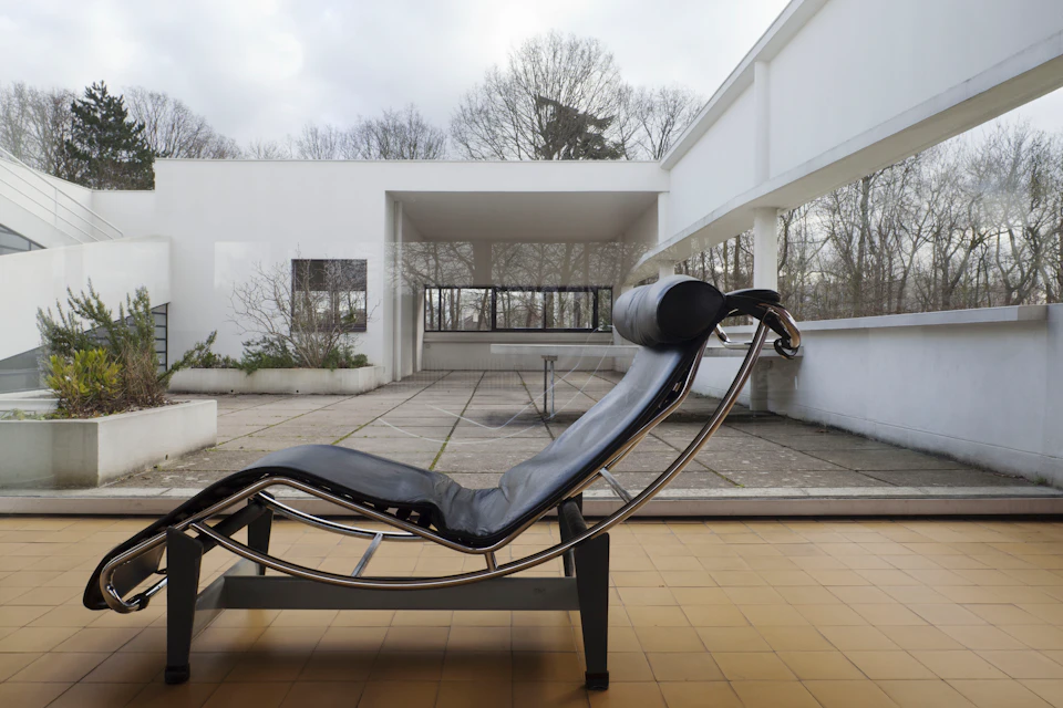 Modern black reclining chair on a terrace with a yellow tiled floor, white walls, and glass doors. Leafless trees in the background.