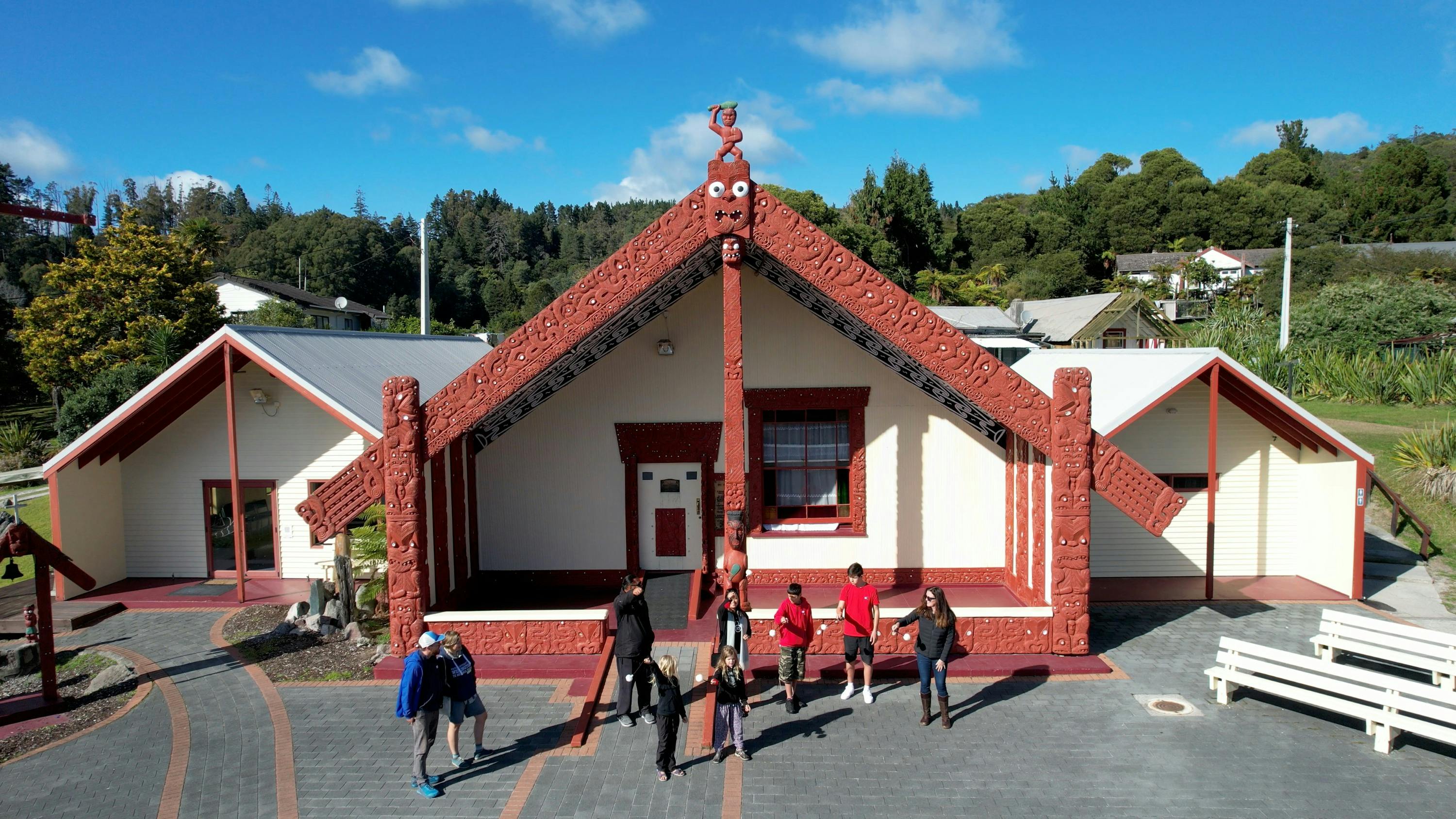Um edifício tradicional de madeira esculpida, com pessoas em frente ao mesmo, rodeado de vegetação sob um céu azul.