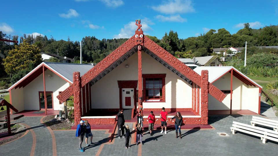 A traditional Māori meeting house with intricate carvings, people standing in front, and trees in the background under a blue sky.