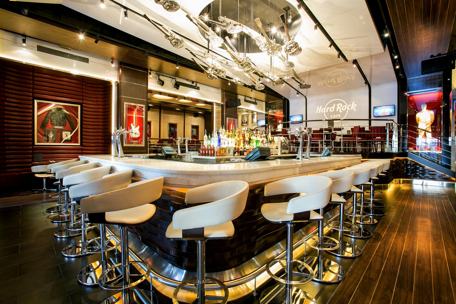 Un bar avec des tabourets blancs, un présentoir central de bouteilles, un décor moderne et le logo "Hard Rock Café" sur le mur.