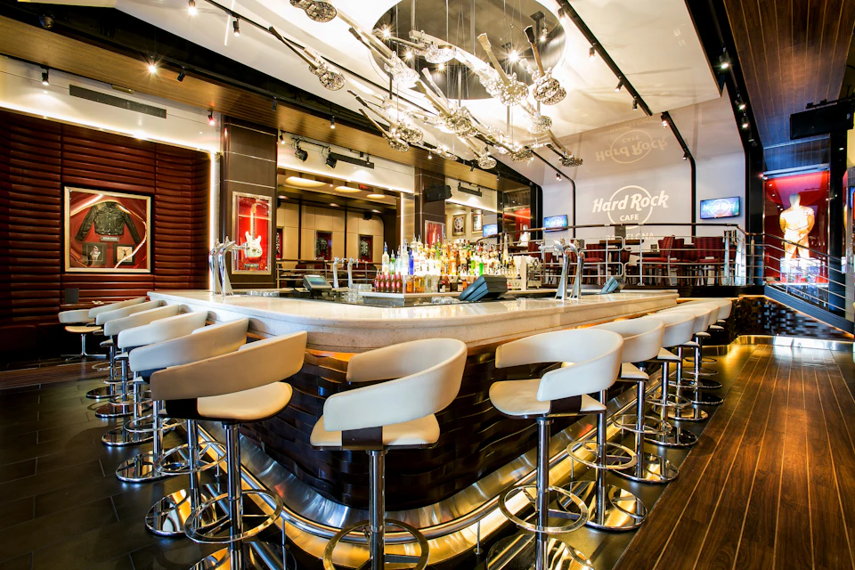 A bar with white stools, centralized display of bottles, modern décor, and "Hard Rock Café" logo on the wall.