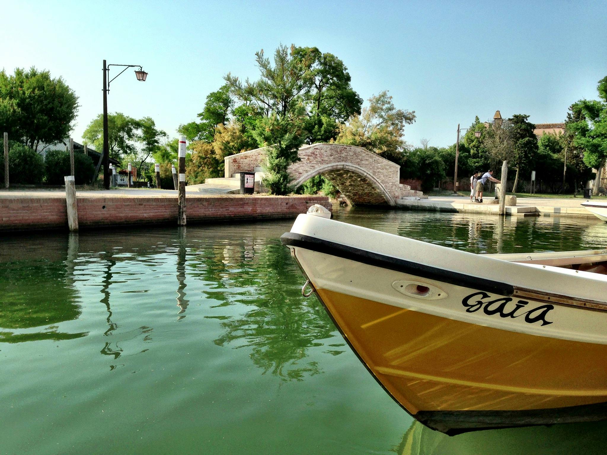 En kanal med en båt som heter "Gaia", en tegelbro, gröna träd, en lyktstolpe och två personer på bryggan.