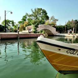 En kanal med en båt som heter "Gaia", en tegelbro, gröna träd, en lyktstolpe och två personer på bryggan.