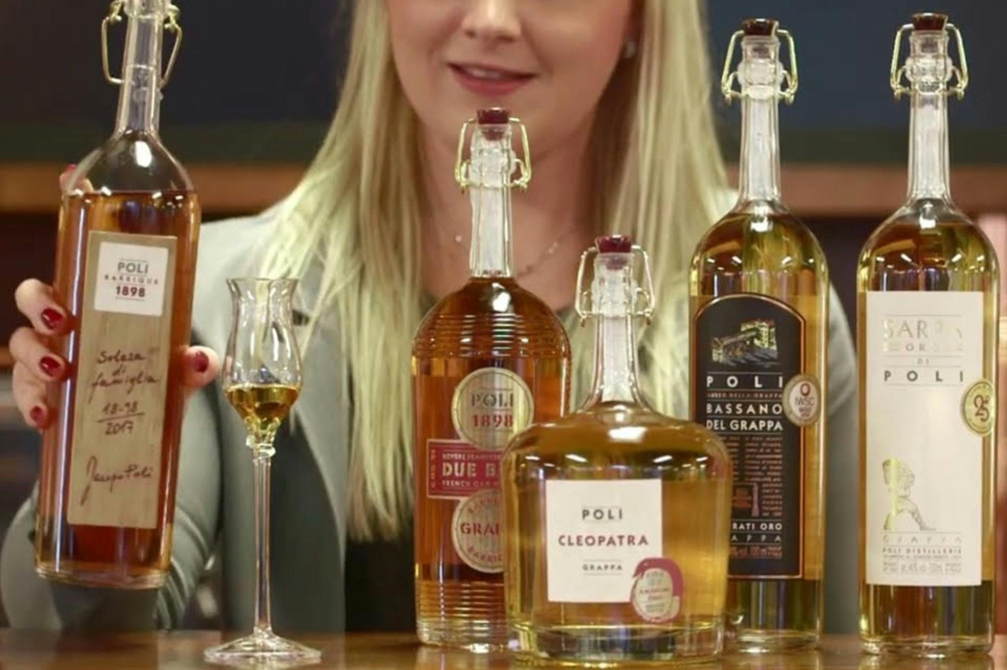 Μια γυναίκα κρατά ένα ποτήρι γκράπα πίσω από τρία μπουκάλια Poli Grappa, συμπεριλαμβανομένων των Poli Cleopatra, Due Barili και Bassano del Grappa.