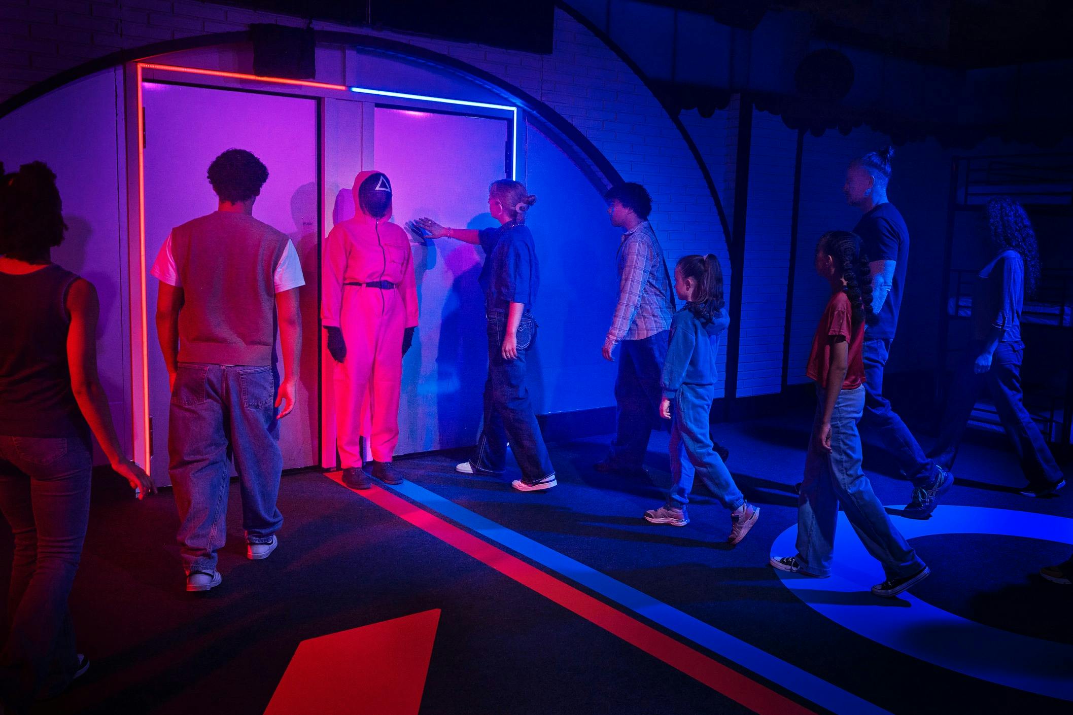 在一间灯光昏暗、有蓝色和红色灯光的房间里，人们排成一排。 一个穿着红色连身裤和面具的人站在门边。