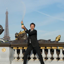 Man i svart kostym sprayar champagne med glädje nära utsmyckade skulpturer, med Eiffeltornet i bakgrunden.