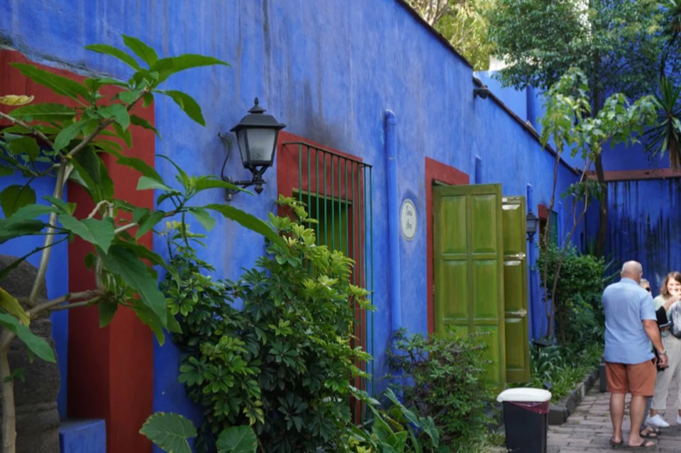 Blue and red building facade with green doors and windows, surrounded by lush foliage and outdoor lanterns.