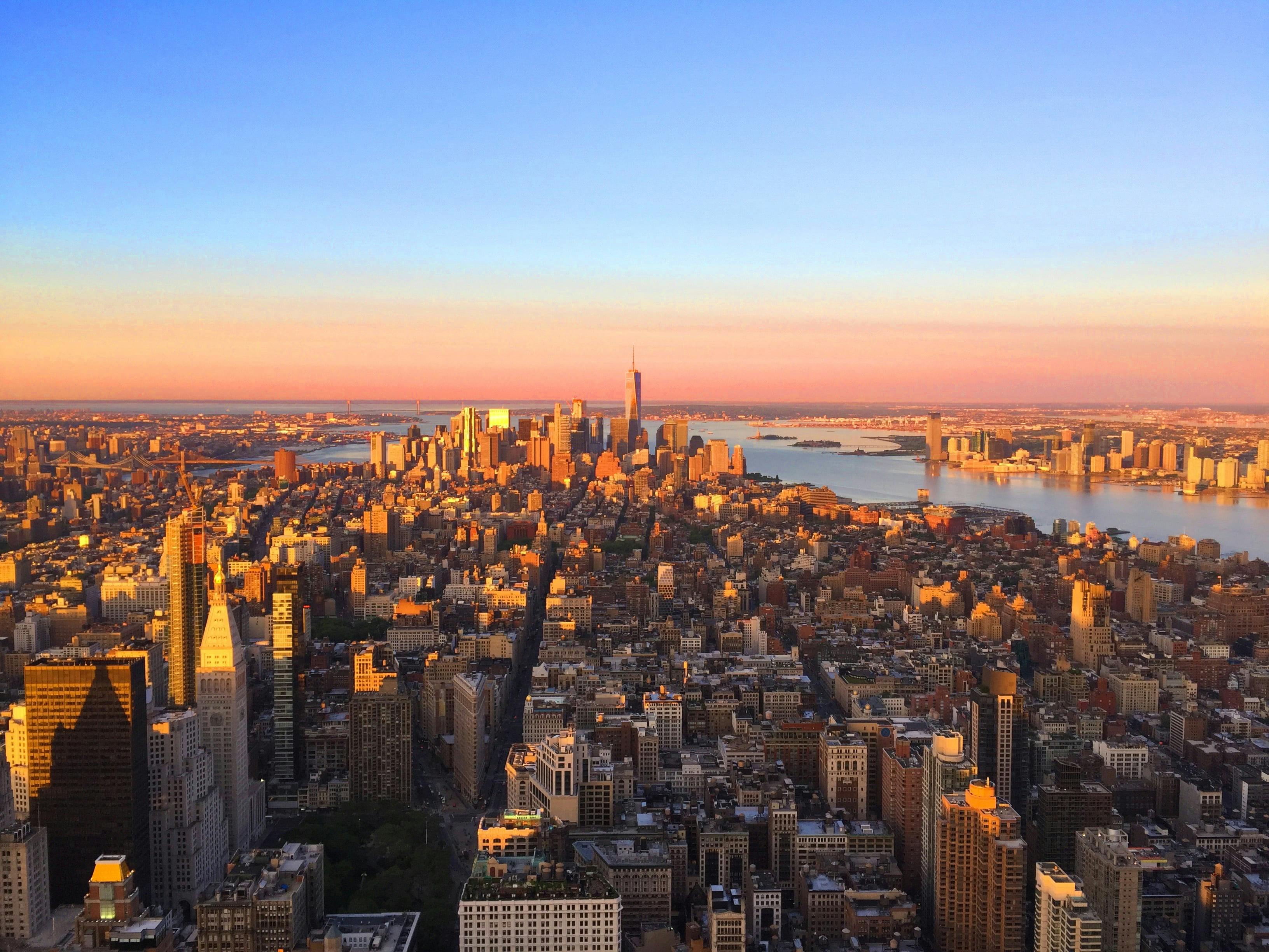 A cityscape view of New York City during sunset, with buildings, the One World Trade Center, and surrounding water visible.