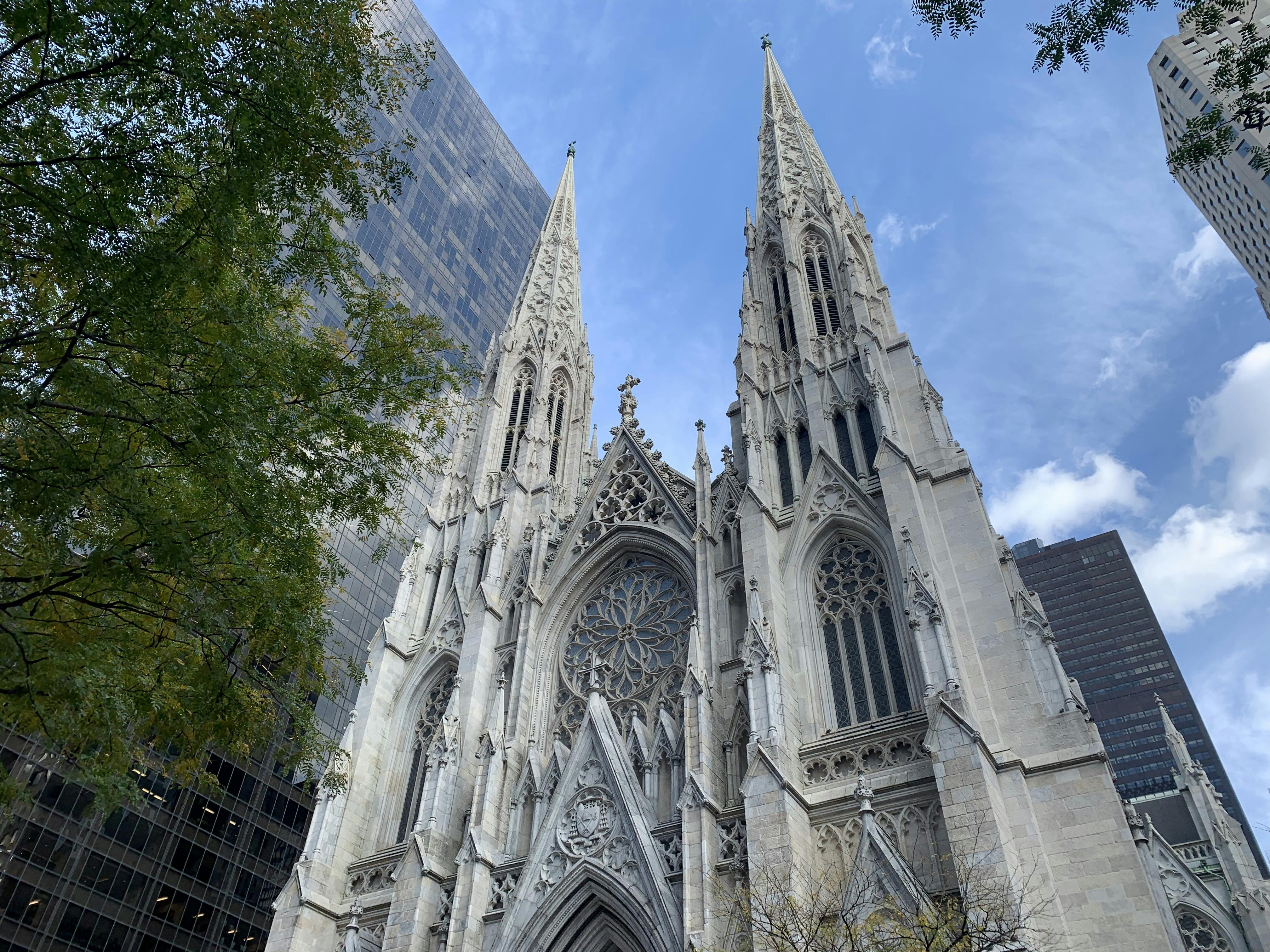 Μυτεροί πύργοι ενός καθεδρικού ναού γοτθικού στιλ με περίτεχνες λεπτομέρειες, πλαισιωμένοι από σύγχρονα κτίρια, δέντρα και έναν εν μέρει συννεφιασμένο ουρανό.